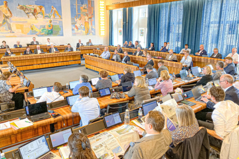 Blick in einen Ratssaal mit vielen Sitzreihen, an Pulten mit Laptops sitzen zahlreiche Menschen während einer Sitzung.