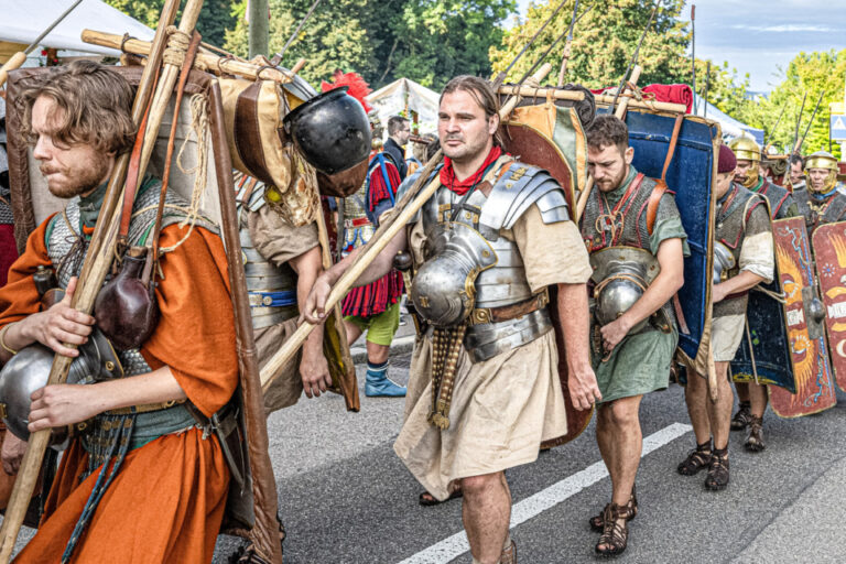 Männer in römischen Legionärskostümen mit Schilden und Speeren marschieren in einer Prozession durch eine Straße.