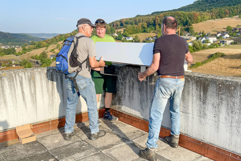 Drei Männer befestigen auf einer Mauer einen weissen Kasten, im Hintergrund hügelige Landschaft.