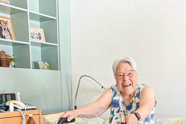 Eine ältere Frau mit weißem Haar lacht, sitzt am Bettrand und hält einen Rollator.