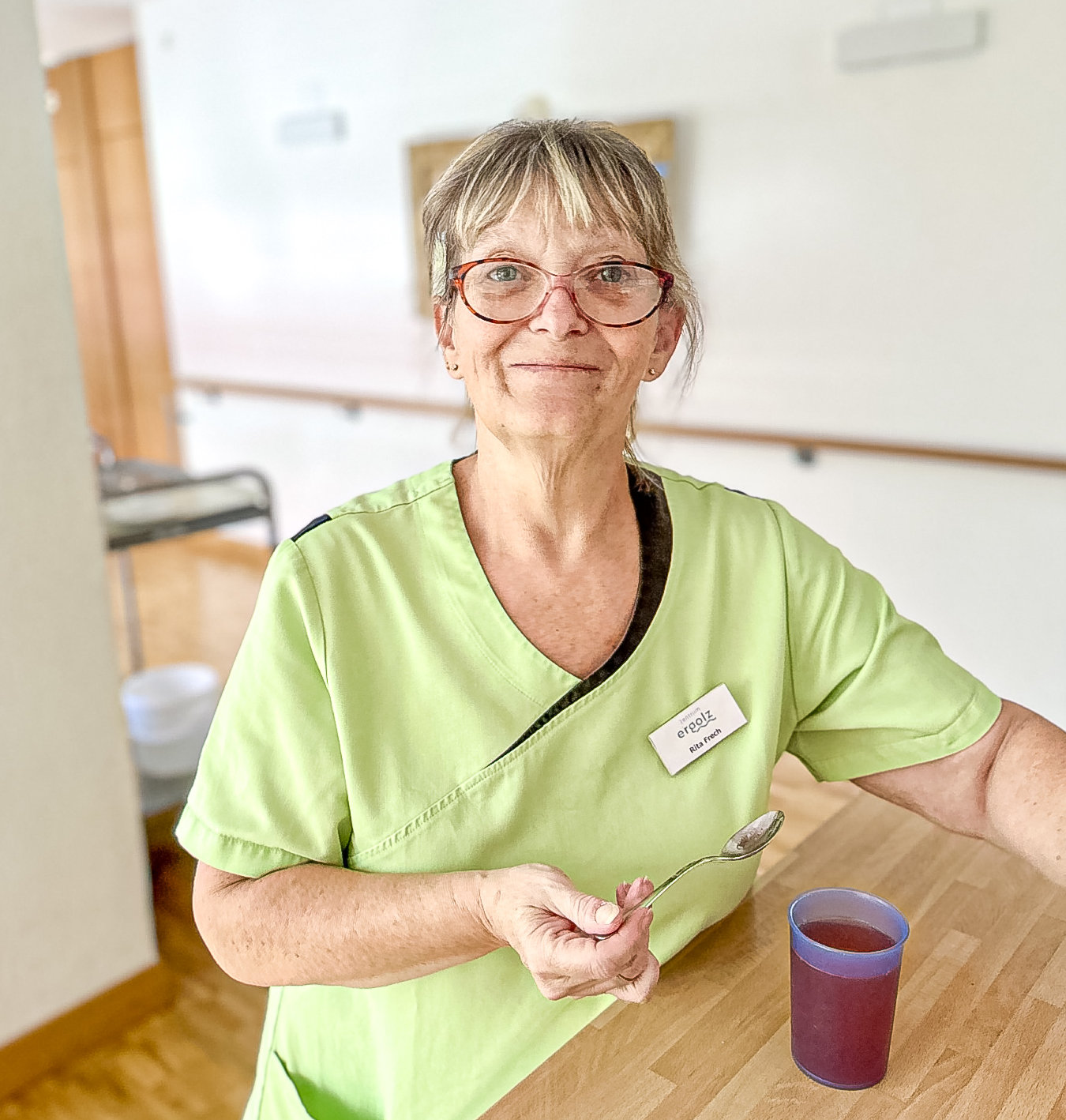 Frau mit Brille und grüner Pflegekleidung sitzt lächelnd an einem Holztisch, hält einen Löffel neben einem Becher.