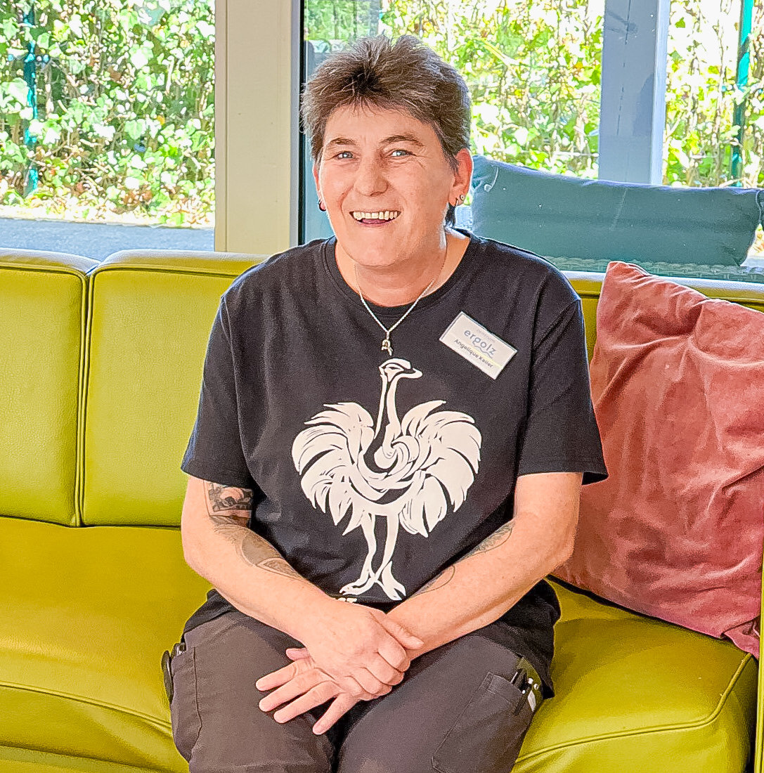 Eine lachende Frau sitzt mit gekreuzten Beinen auf einem gelbgrünen Sofa, sie trägt ein T-Shirt mit Vogelmotiv.