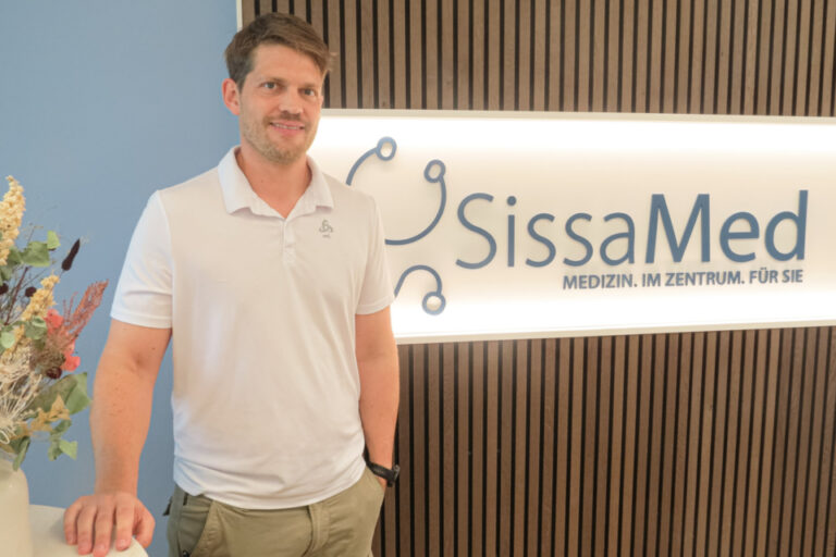 Ein lächelnder Mann im weissen Poloshirt steht vor einem beleuchteten Praxisschild mit der Aufschrift SissaMed.