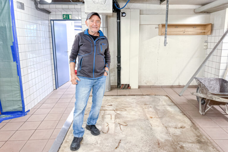 Ein älterer Mann mit Cap steht lächelnd in einem Kellerraum vor einer ausgehobenen Bodenfläche.