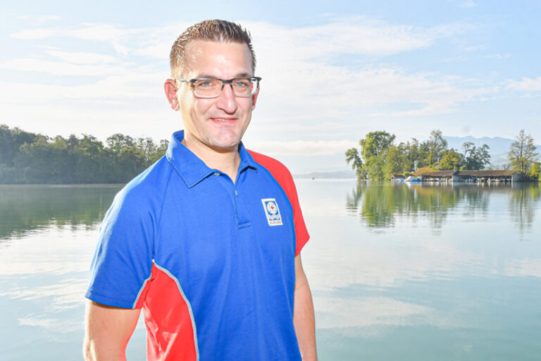 Ein Mann mit Brille und Polo-Shirt lächelt vor einem ruhigen See mit bewaldeten Ufern.