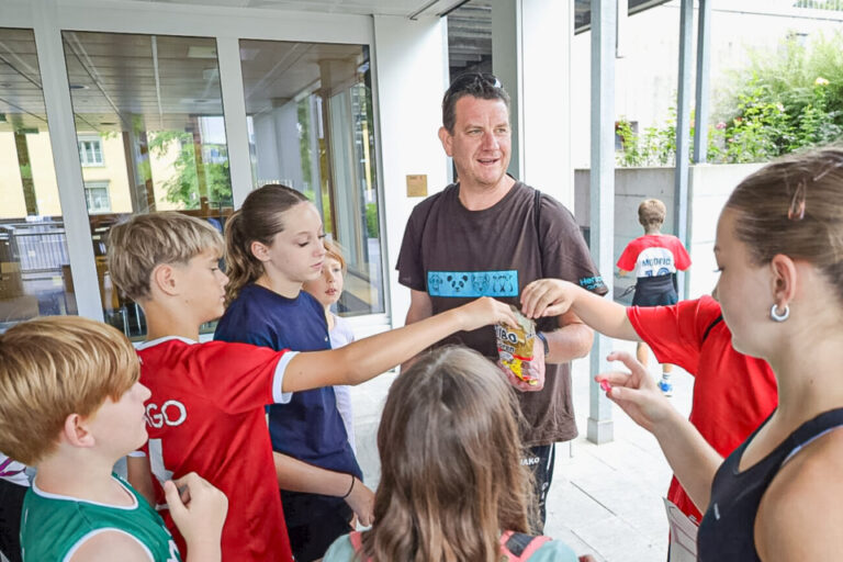 Ein Mann steht mit mehreren Kindern im Kreis vor einem Gebäudeeingang und verteilt kleine Gegenstände.