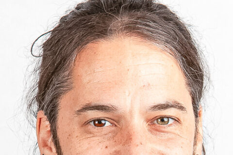 Ein lächelnder Mann mit zusammengebundenen Haaren und Bart trägt ein geblümtes Hemd.