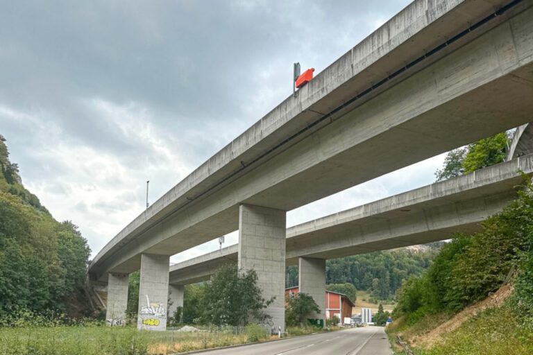 Mehrstöckige Betonautobahnbrücke überspannt eine leere Landstrasse zwischen bewaldeten Hügeln.