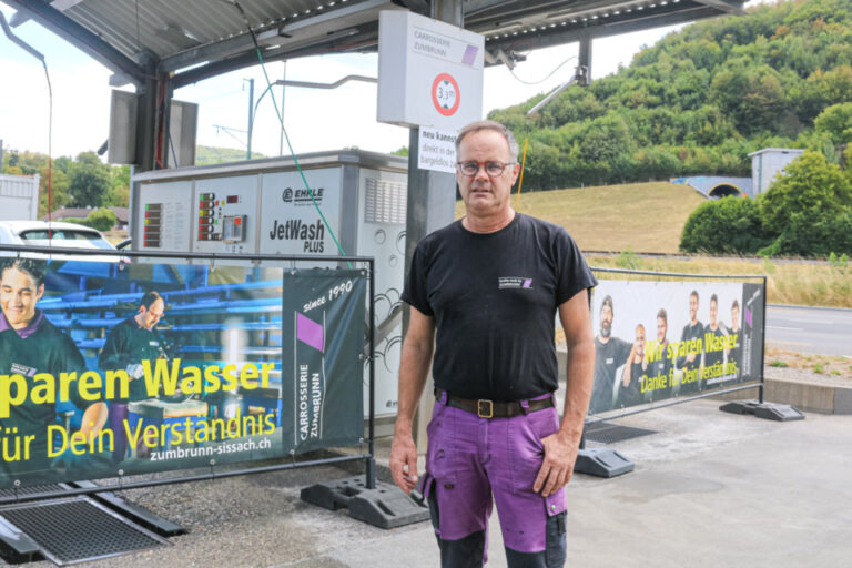 Ein Mann mit Brille und schwarzem T-Shirt steht vor einer Autowaschanlage mit Werbebannern.
