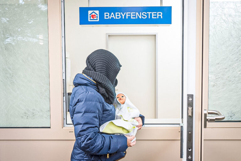 Eine vermummte Person hält ein Baby vor einer Tür mit der Aufschrift „Babyfenster“.