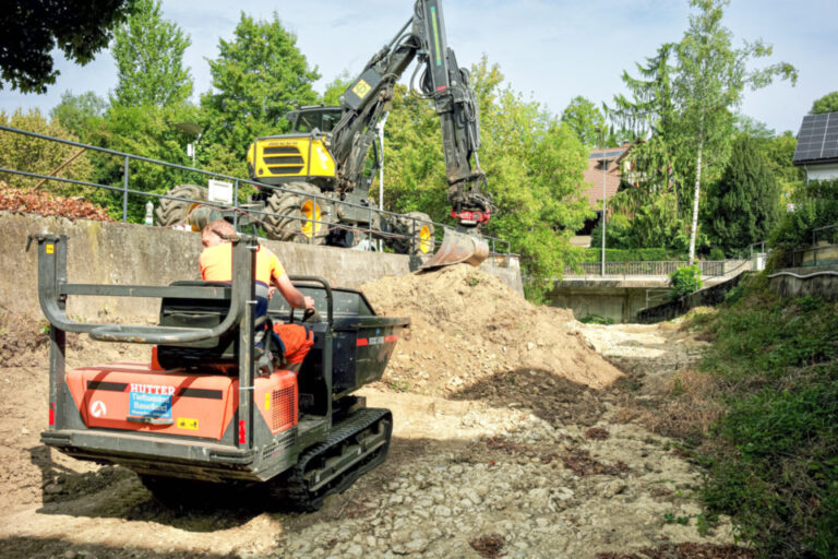 Ein Bauarbeiter sitzt auf einem Kettendumper neben einem Bagger auf einer Baustelle mit Erdaushub.