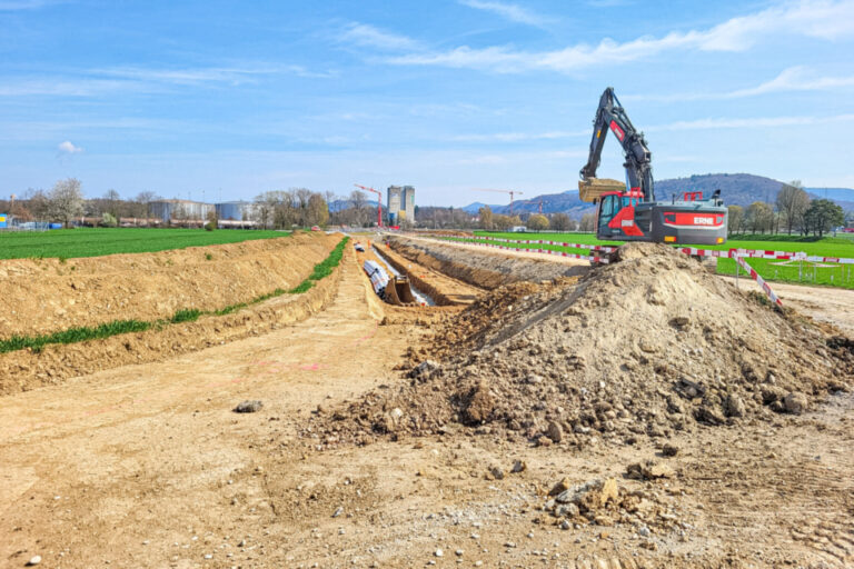 Bagger gräbt einen langen Erdgraben mit verlegtem Rohr neben einem Feld, Hügel im Hintergrund.