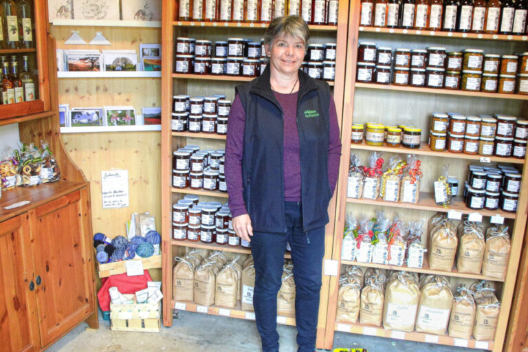 Eine Frau steht in einer Weste in einem Hofladen vor Regalen mit Marmeladengläsern und Gebäcksäcken.