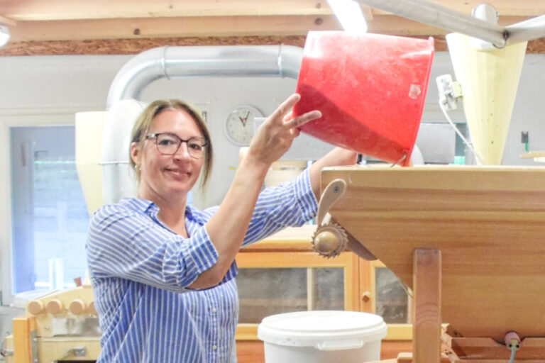 Eine Frau in gestreifter Bluse giesst Getreide aus einem roten Eimer in eine hölzerne Steinmühle.