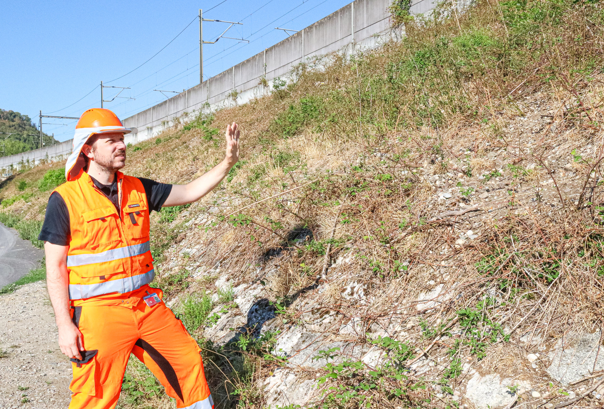 Ein Mann in oranger Arbeitskleidung und Helm steht an einer steilen, bewachsenen Böschung und deutet mit der Hand darauf.