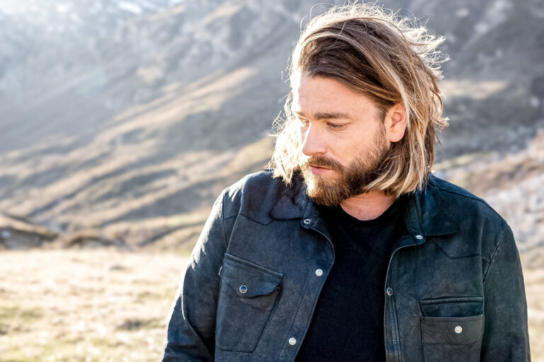 Ein bärtiger Mann mit schulterlangen Haaren blickt zur Seite, im Hintergrund eine Berglandschaft.