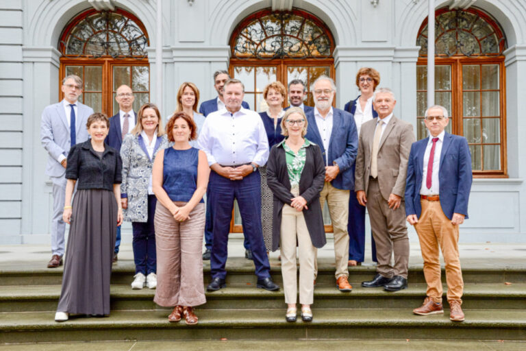 Eine Gruppe von Männern und Frauen in zwei Reihen posiert auf einer Steintreppe vor einem historischen Gebäude.