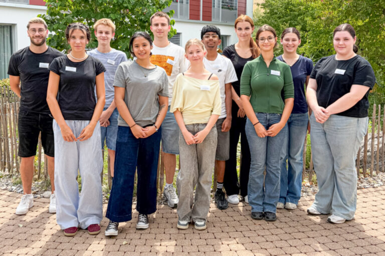 Elf junge Erwachsene stehen nebeneinander im Freien vor einem Gebäude mit Bäumen im Hintergrund.