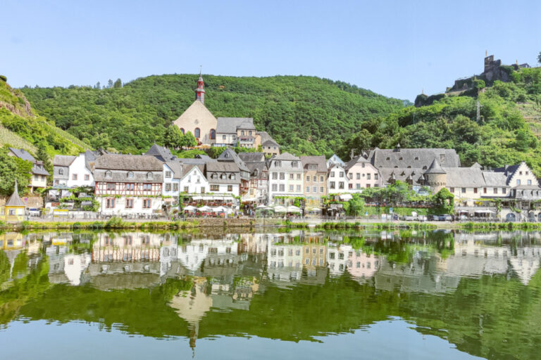 Ein Weinort mit Fachwerkhäusern und Kirche am Flussufer, im Hintergrund eine Burgruine auf bewaldetem Hügel.