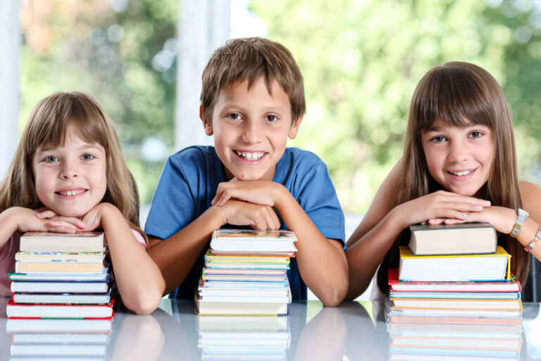 Drei lächelnde Kinder stützen sich auf Bücherstapel und blicken in die Kamera.