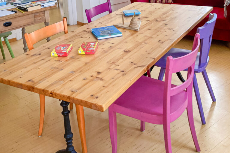 Heller Raum mit Holztisch, bunten Stühlen und rotem Sofa, Spiele liegen auf dem Tisch.