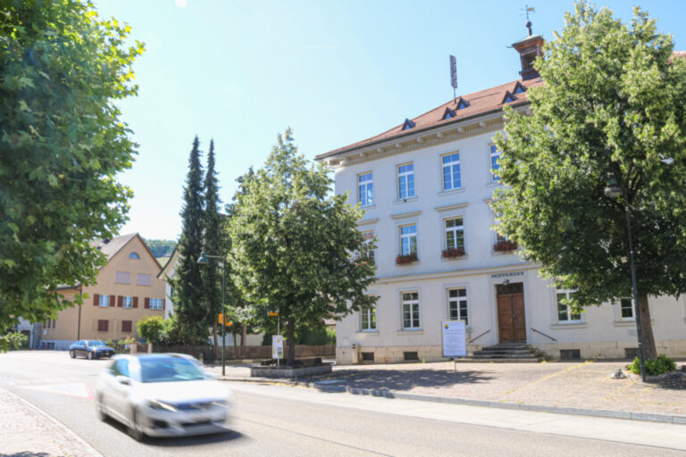 Historisches, weiss verputztes Gebäude mit rotem Ziegeldach hinter Bäumen, davor fährt ein Auto vorbei.