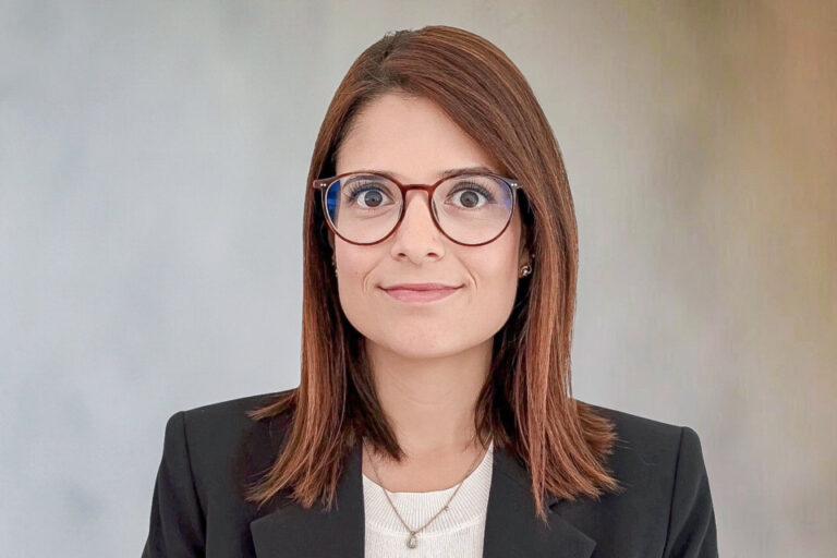 Frau mit schulterlangem braunem Haar und Brille im dunklen Blazer blickt in die Kamera.
