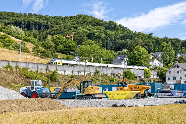 Baustelle mit Bagger und Radlader vor einer Böschung, im Hintergrund fährt ein Zug über eine Brücke.
