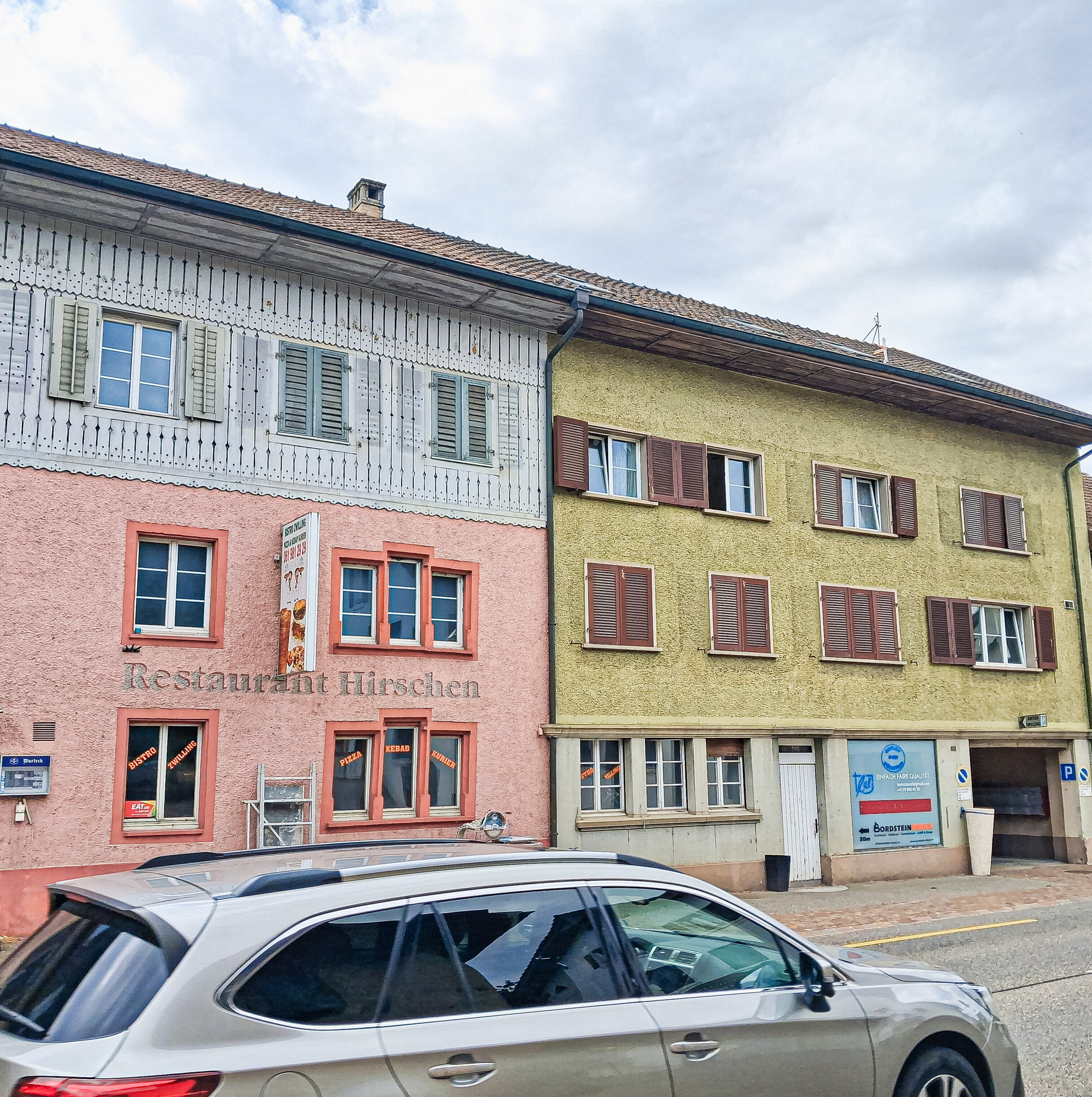 Rosa und gelb gestrichene historische Häuserzeile an einer Strasse mit parkiertem Auto davor.