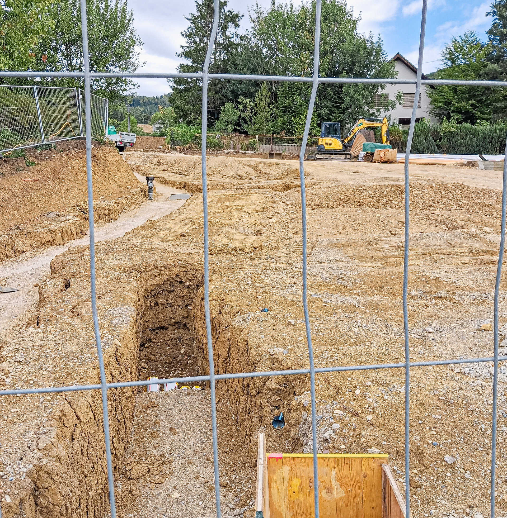 Hinter einem Bauzaun verläuft ein ausgehobener Graben durch eine aufgewühlte Baustelle mit Bagger im Hintergrund.