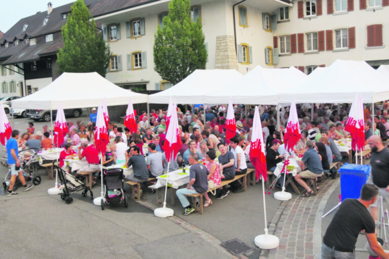 Menschen sitzen an langen Tischen unter weissen Zelten mit roten Fahnen bei einem Dorffest.