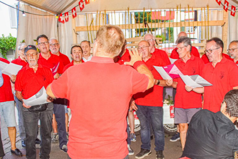 Männerchor in roten Shirts singt unter einem Zeltdach, ein Dirigent in Rot leitet vor ihnen.
