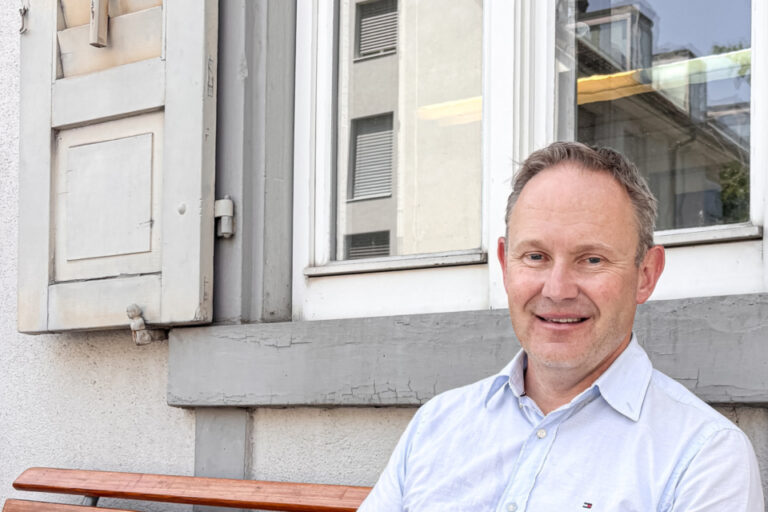 Ein lächelnder Mann in hellblauem Hemd sitzt auf einer Holzbank vor einem Fenster.
