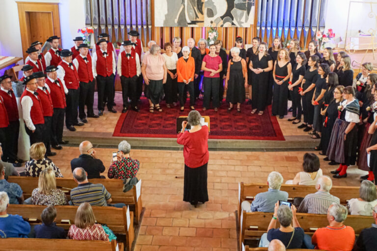 Mehrere Chöre in unterschiedlichen Trachten und Uniformen singen gemeinsam vor einer Orgel in einer Kirche, davor eine Dirigentin und Zuschauer in Bankreihen.