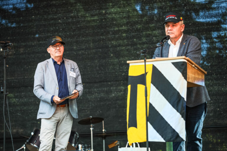 Zwei Männer mit Kappen auf einer Bühne, einer spricht am Rednerpult mit Wappen-Banner.