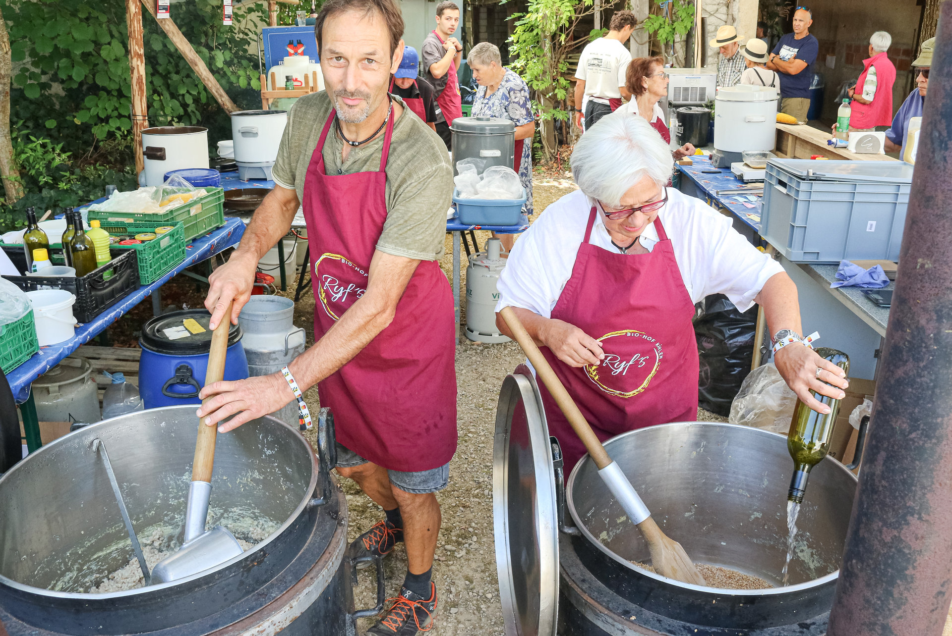 Zwei Personen in weinroten Schürzen rühren mit Holzkellen in grossen Metallbottichen im Freien.