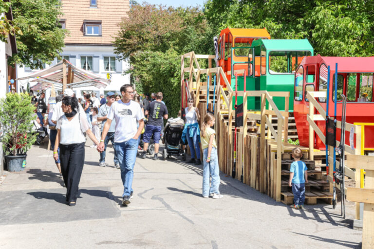 Ein Paar geht händchenhaltend über einen Platz, im Hintergrund Kinder vor bunten Holzspielgeräten.