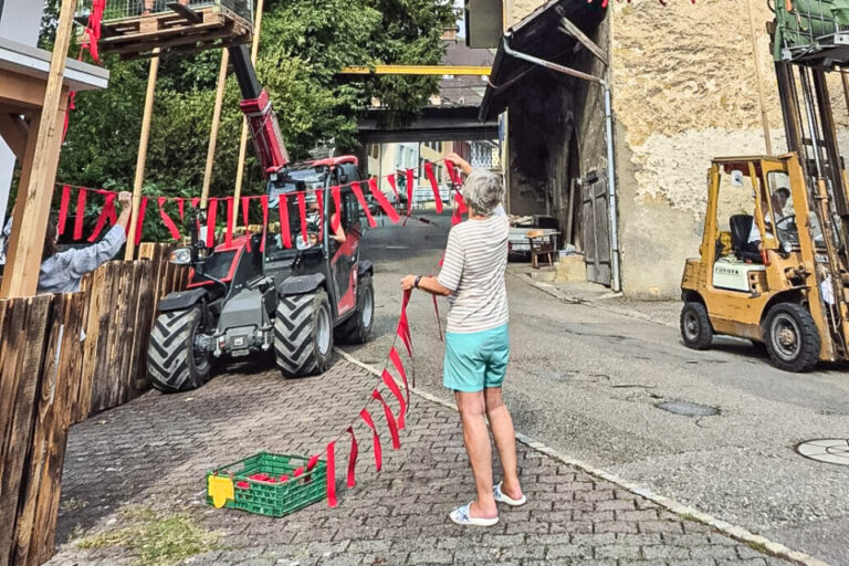 Eine Person spannt rote Papiergirlanden über eine Dorfstrasse, im Hintergrund Hubarbeitsbühnen und Häuser.