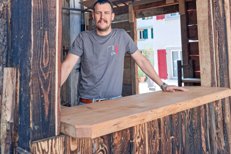 Ein Mann mit Bart lehnt an einem Holztresen in einer rustikalen Hütte aus Holzbalken.