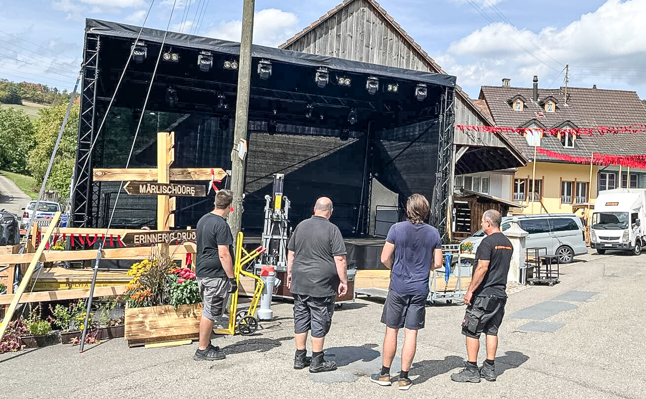 Drei Männer stehen vor einer schwarzen Konzertbühne, im Hintergrund Bauernhäuser mit roter Fahnendekoration.