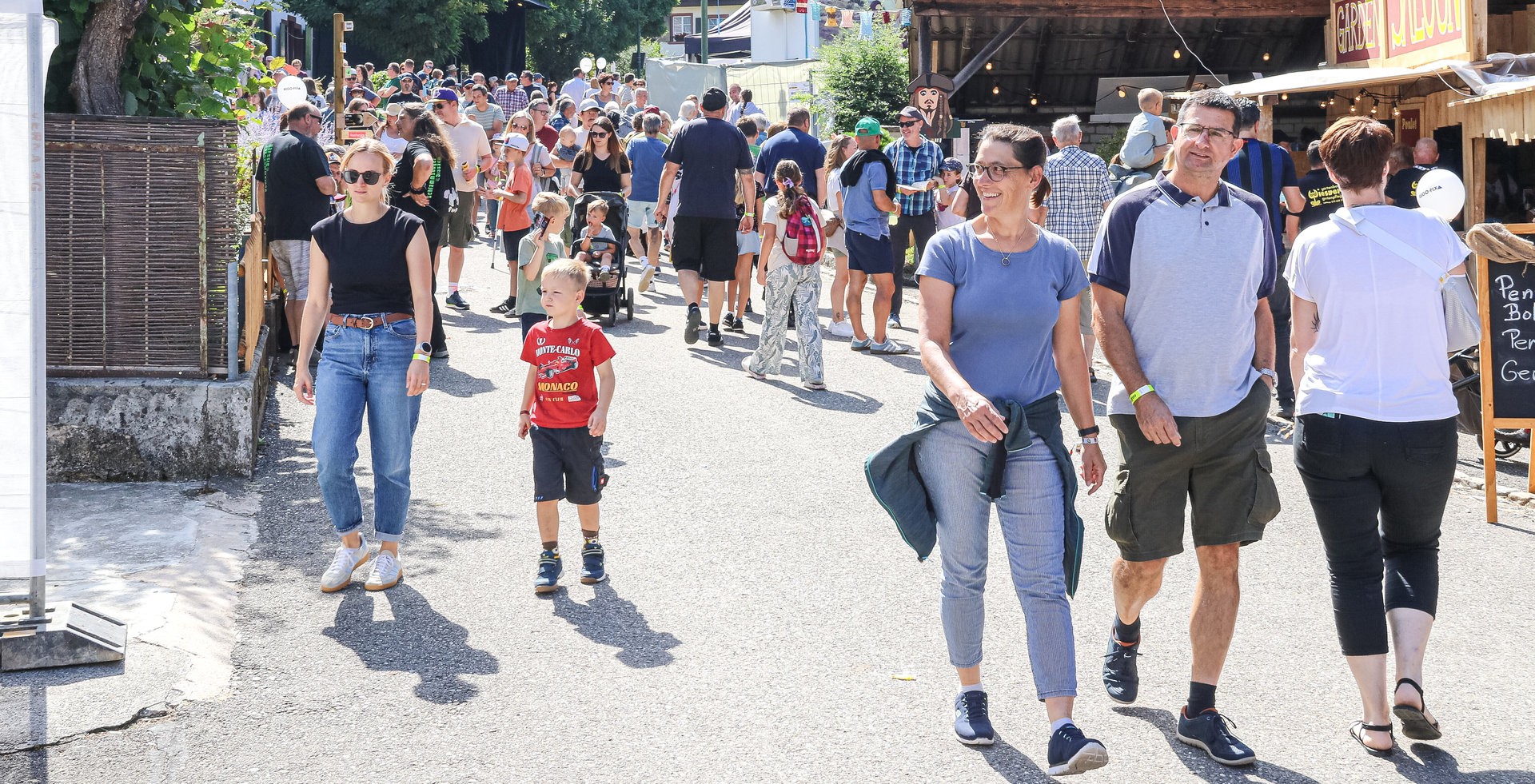 Menschenmenge schlendert an einem sonnigen Tag durch eine belebte Dorfstrasse mit Marktständen.