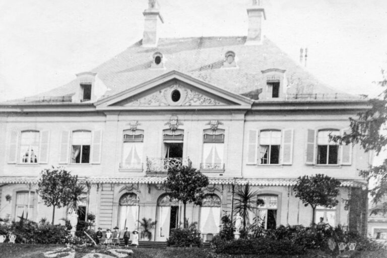 Historische Aufnahme einer herrschaftlichen Villa mit Türmchen, Fensterläden und Garten, davor stehen mehrere Personen.