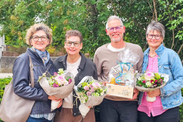 Drei Frauen und ein Mann stehen lächelnd nebeneinander vor Sträuchern, sie halten Blumensträusse und ein Geschenkkorb.