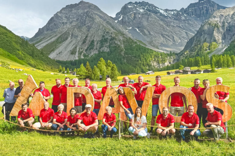 Grosse Gruppe in roten Poloshirts posiert um hölzerne Buchstaben "DAVOS" in einem grünen Alpental vor Bergpanorama.