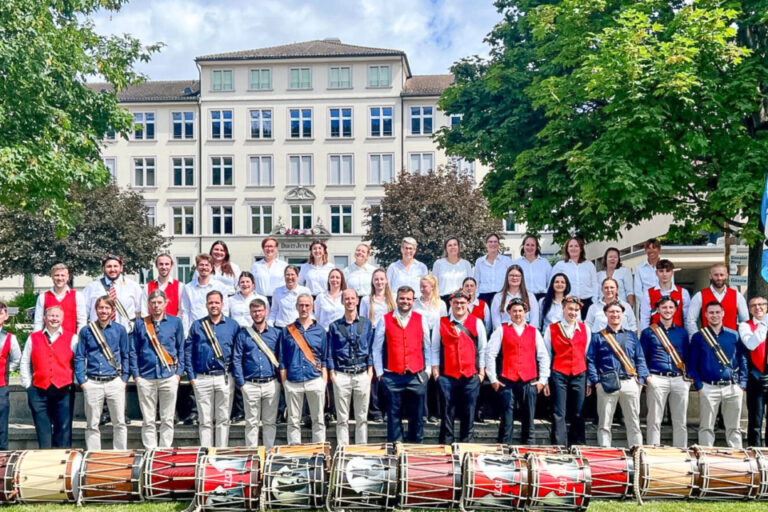 Gruppenfoto einer Trommler- und Chorformation in Trachten vor einem Gebäude, mit Trommeln auf dem Rasen davor.