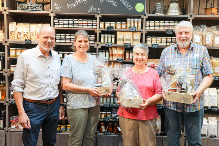 Vier lächelnde Personen stehen mit Geschenkkörben in einem Laden vor Regalen mit Lebensmitteln.