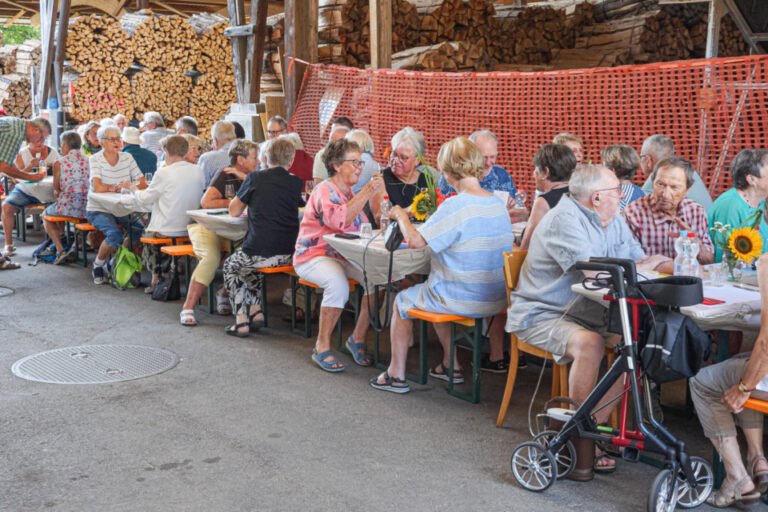 Ältere Menschen sitzen an langen Tischen im Freien unter einem Holzschuppen und unterhalten sich beim Essen.