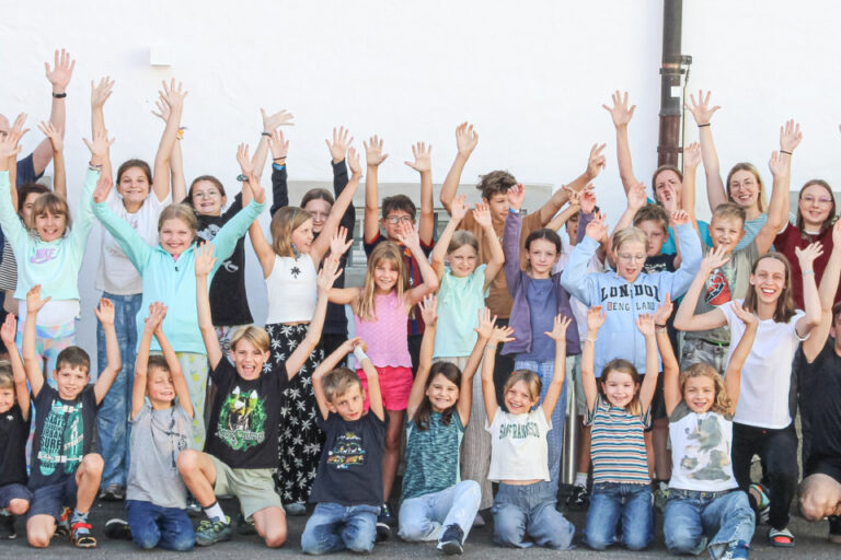 Eine grosse Gruppe von Kindern und Erwachsenen steht jubelnd mit erhobenen Armen vor einer Hauswand.