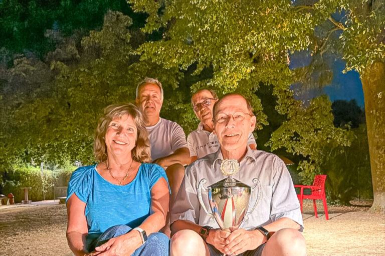 Vier lächelnde Personen sitzen abends im Freien auf einer Bank, der Mann vorne rechts hält einen Pokal.