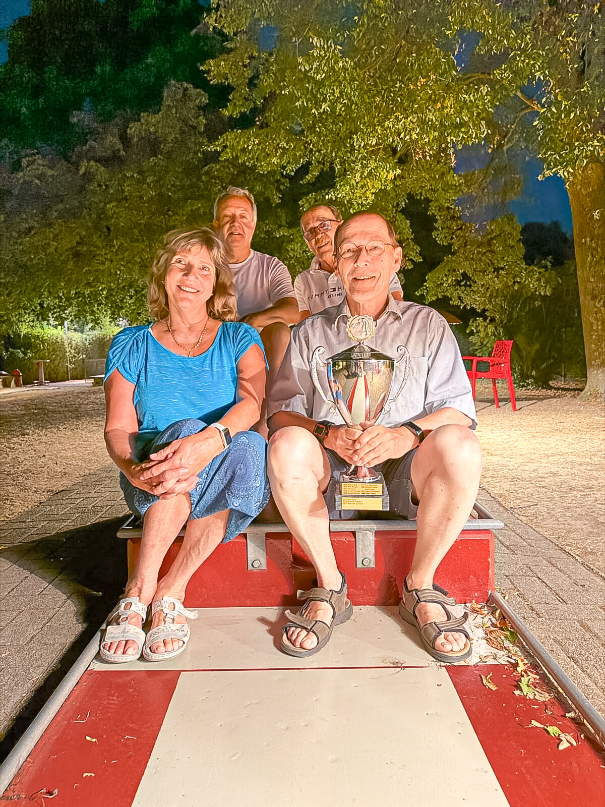 Vier lächelnde Personen sitzen abends im Freien auf einer Bank, der Mann vorne rechts hält einen Pokal.